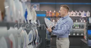 一个穿衬衫的年轻英俊的男人在一家消费电子商店里选择熨斗。 学士